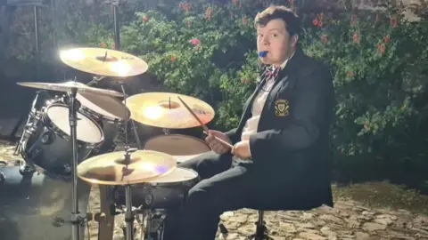 Harry Chambers A 20-year-old man called Harry Chambers sits by a drum kit. He is wearing a suit with a Union Flag bow tie on. He has a blue whistle in his mouth and drumsticks in his hands.