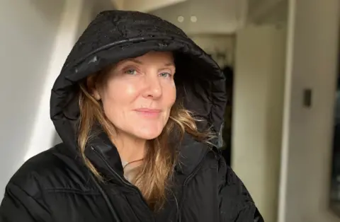 BBC/Harry Low A woman with long, light coloured hair in a black coat with the hood up is interviewed in a car park in the rain. There are cars parked behind her. 