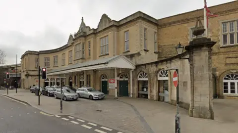 A view of Bath Spa train station on Dorchester Street. The station is a beige building and taxis are parked outside.