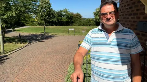 Resident Mark Wood photographed in front of his house at Ash Way on a sunny day. Behind him, there is a green space and a sign with the road's name