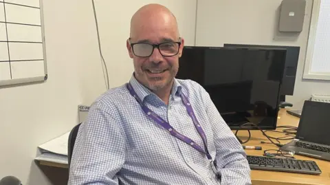 A bald man sitting in an office chair at a desk, he is wearing a blue and white checked shirt and has a purple lanyard around his neck. He is wearing a pair of black framed glasses, his right glasses' lens is clouded whilst the left lens is transparent. To the man's right is a support stick which he is not holding.