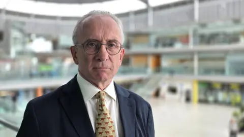 Paul Moseley/BBC David Bick is wearing a dark blue suit jacket over a white shirt. He has a gold and  red patterned tie.