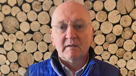 Rowland Rees Evans standing in front of a pile of logs. He is wearing a blue jacket and has glasses on.