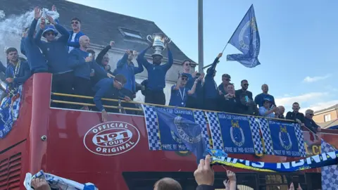 Dungannon Swifts players wearing blue tops and black trousers and many wearing sunglasses parade on top of a red open-top bus with team flags draped over the sides 