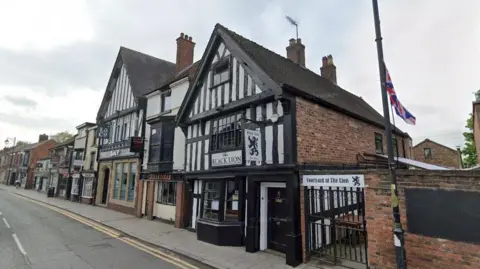 Google A black and white building fronted in Nantwich, in a row of buildings stretching alongside a road with a pavement running between the building and road. There is a gated courtyard to the side of the building.