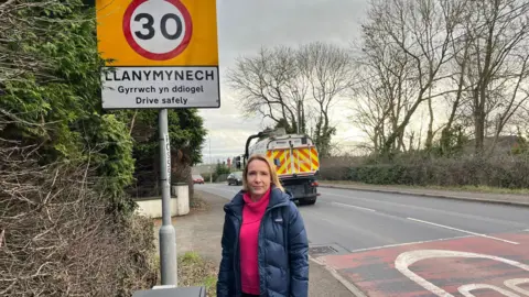 A woman wearing a pink hoodie and a navy blue puffer coat stands next to a 30mph road sign reading Llanymynech with a road behind her.