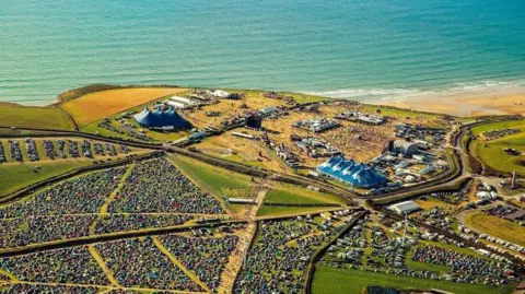 An overhead photo showing the scale of the festival site, with tents in fields. stages, arenas and a beach in the background