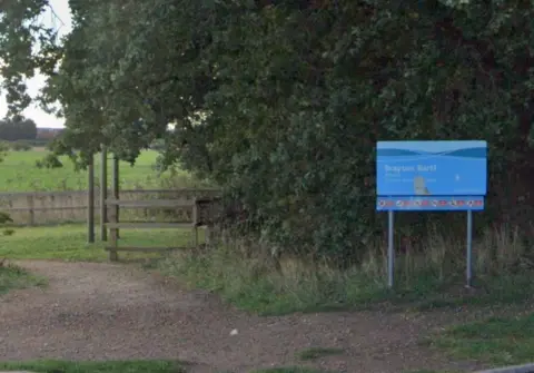 A blue sign at the entrance to a nature reserve with a rough path leading to the left of the picture through trees to an open field.