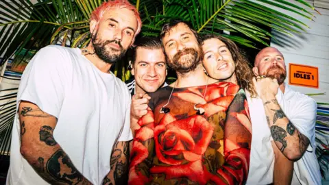 A press shot of the Idles smiling to camera, hugging each other and wearing white t-shirts and tropical style clothing with a bamboo plant in the background