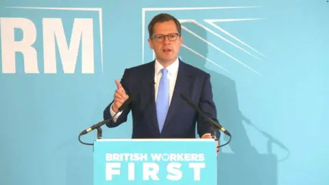PA Media Robert Jenrick MP in a white shirt and navy blue suit with a light blue tie. He is standing behind a lectern and in front of a Reform background in the turquoise colours of the party.