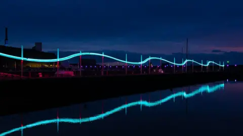 Deanne Shallcross An image of the Wave light installation illuminated in blue at night at Whitehaven Harbour. It is installed on a walkway leading out into the middle of the harbour and is in the shape of a stylised wave.