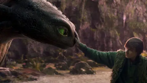 A boy with swept over to the side hair, wears a green outfit, fluffy waistcoat puts his hand out and touches the nose of an animated dragon which has green eyes.