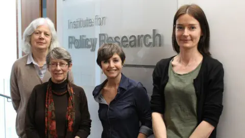 University of Bath The UoB team looking into Universal Credit, from left to right: Professor Jane Millar, Fran Bennett, Rita Griffiths and Marsha Wood. They are all wearing shirts with blouses, and one is wearing an orange and brown scarf. They are leaning on a wall where a big glass sign reads 'Institute for Policy Research'.