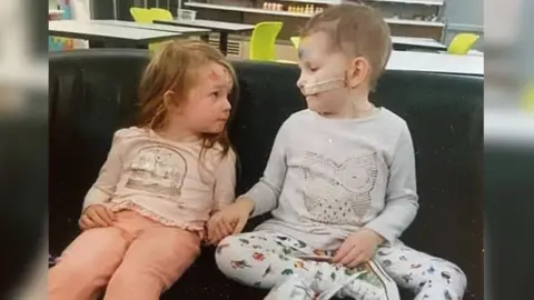 Sam Tucker Two little girls are sat on a chair holding hands. They are looking directly at each other. They are both around three or four years-old and wearing leggings and a long sleeve top. 
The child on the left has brown hair, the child on the right has a feeding tube in her nose and very little hair. 