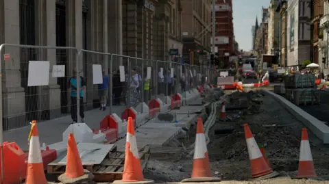 Roadworks along a city centre street with traffic cones, safety barriers and fenced-off excavations. Pedestrians walk along a temporary pavement beside construction works, with buildings and traffic visible in the background.