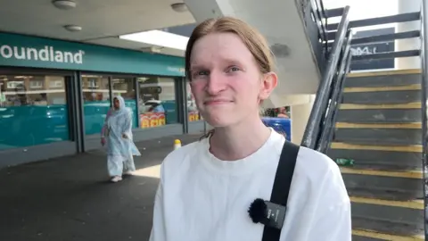 A young white man with reddish blonde hair tied back smiles at the camera. He is in the street, and wears a white top. A branch of Poundland is pictured behind him, as is a concrete flight of stairs.