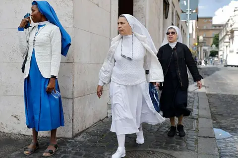 MARIO TAMA/GETTY IMAGES A Nigerian reverend sister wearing a blue habit on the phone as two other sisters wearing black and white habits walk by on the streets of Rome. 