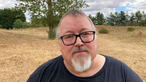 Jason Kernohan stood in Burlish Meadows wearing a black t-shirt and black glasses. Jason also has a white goatee beard and short white hair.