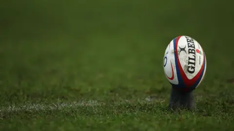 Getty Images A rugby pitch showing signs of degradation with bits of mud appearing through the grass. There is a white line marking on the pitch. A gilbert rugby sits on a tee on the right of the image.