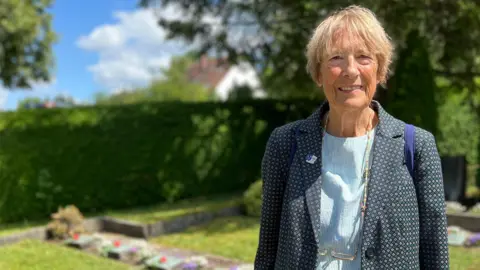 Lola stands in a well-maintained graveyard, wearing a dark blazer over a light blue shirt and a lanyard. The background includes green hedges, trees, flower beds, and a partly cloudy sky with blue patches