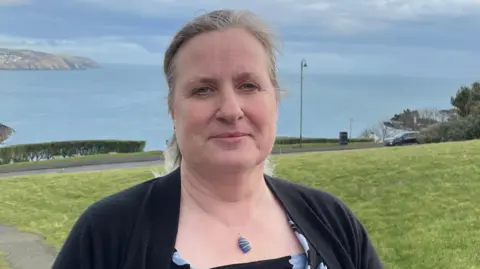 Michelle Haywood, who is wearing a dark cardigan over a blue and black flowery blouse, and a necklace. She has fair hair which has been tied back and there is a large patch of green grass in the background in front of Douglas Bay.