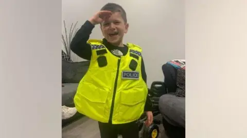 Northumbria Police Archie York is smiling as he wears a high visibility police vest. He is holding his right hand next to his forehead for a salute. He has short brown hair.