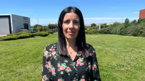 Rocio Cifuentes stands in a field looking at the camera. She has long dark hair and is wearing a floral blouse.