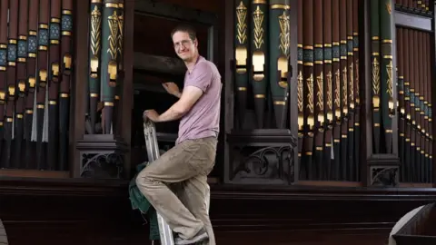 A man in cream jeans and a pink T shirt is pictured climbing a ladder and smiling at the camera before climbing inside an historic pipe organ to start restoration work. 