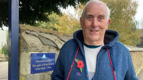 An older man standing outside a memorial garden with a poppy on his blue fleece jacket and the red string holding his tray of poppies around his neck