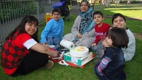 Sabah Usmani and her five children smile at the camera as they sit around a birthday cake in a park. Dr Usmani is wearing a blue headscarf and grey cardigan. 