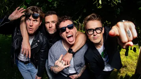 McFly, four band members, in a row. Dougie, is to the left, with sunglasses raised, with his hand on his glasses, he is wearing blue jeans, a white T-shirt and black jacket, Harry,  has his around round two men, Fanny, has two fingers raised, is wearing blue sunglasses, a blue top and his mouth open, Tom is pointing his finger, and is wearing a T-shirt and jacket. 