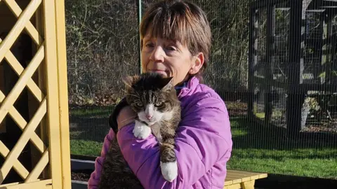 Meryl Marshall is wearing a purple coat and has short brown hair. She is holding a big tabby cat called Fred who has white paws, close to her body. 