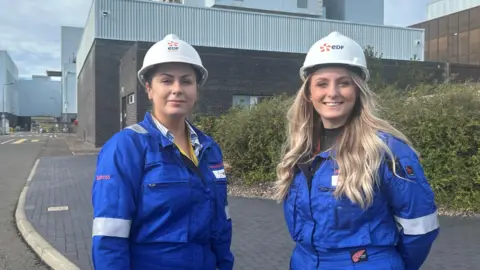 EDF Energy Two women wearing blue coveralls and white helmets standing in front of the main turbine hall at Torness power station.