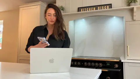 A woman with long dark hair is looking at a laptop on a kitchen worktop. She is holding a mobile phone in one hand.