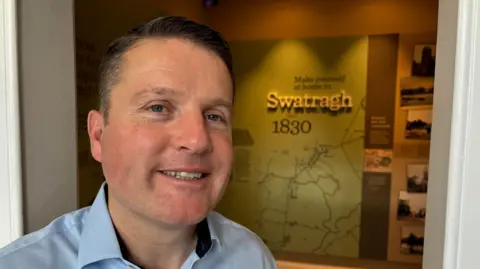 Dermot Friel wearing a blue shirt, standing in front of a wall with 'Make yourself at home in Swatragh' written on it. 