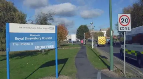 BBC An entrance road for the Royal Shrewsbury Hospital. On the right, ambulances can be seen passing a 20 mph speed limit sign. On the left is a blue and white sign saying "Welcome to Royal Shrewsbury Hospital".