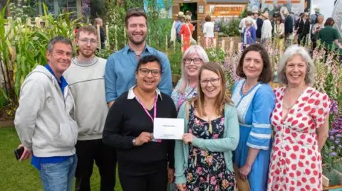 J.Garget/University of Cambridge A group of eight people huddled in front of flowers, with two women at the front holding their award. They are standing together as part of a group and are all smiling at the camera in front of their exhibition.