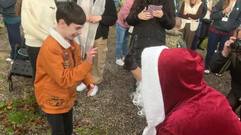 A boy in an orange jacket about to high-five Santa outside, who is kneeling down