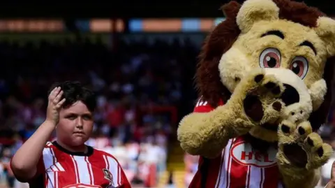 Family photo Oliver stood to the left of a lion mascot. They are both wearing a red and white Exeter City FC football shirt. The lion mascot has its paws in front of its face and Oliver has his left arm raised.