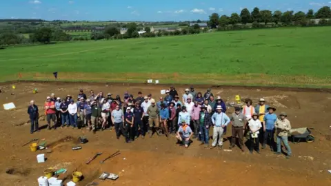 University of Leicester A large group of people, standing in a field, but archaeology items. Grass is behind them, buildings and trees. 