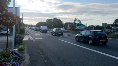 Cars are lined up on a double-carriageway road which has been reduced to one lane, with orange and white cones blocking off the other lane