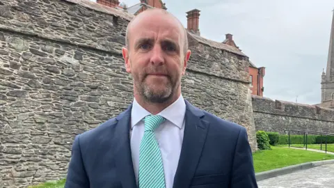 Mark H Durkan is wearing a suit and tie. His blazer is navy, shirt is white and his tie is green and white stripes. He has facial hair. Behind him are the walls of Derry and a red brick building. There is a paved grey path and green grass between him and the walls. In the distance we can see a large stone church tower.