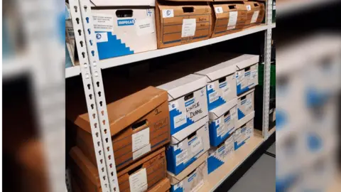 Avon and Somerset Police Stacks of boxes on a metal shelf. The words "Louisa Dunne" are visible on some of the boxes.