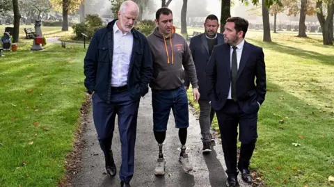 Bernard Platt Four men walk along a concrete path in an area of parkland with a plot of land lined with gravestones in the backdrop. The man in the middle is wearing a pair of denim shorts over two prosthetic limbs.