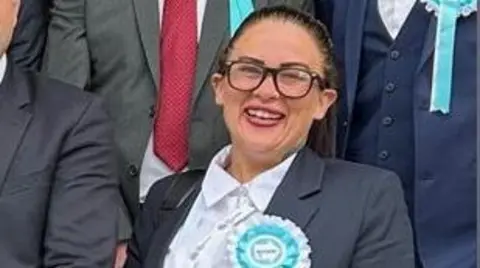 Kathryn Shaw stands next to council leader, Mark Arnull. They both wear suits and Shaw has a pale blue rosette indicating Reform UK on her jacket lapel. Other members of the Reform Party are stood behind them but you cannot see their faces.
