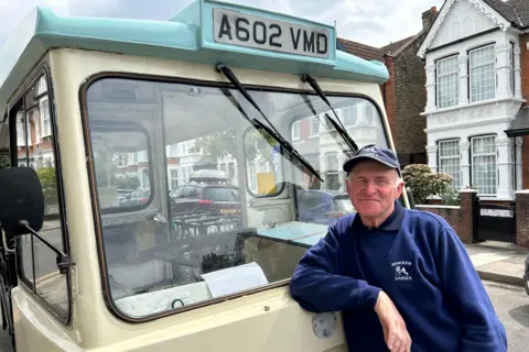 BBC/Harry Low A man wearing a blue Parker Dairies baseball hat and jumper with a cow logo on leans on the front of his milk cart and smiles at the camera. There are crates visible through the front window on the back of the cart. There are residential homes in the background.