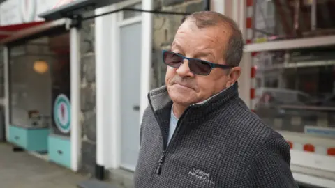 Phil, a man in a dark jumper wearing sunglasses and standing in front of a butcher shop waiting for the bus. He has short dark hair. 