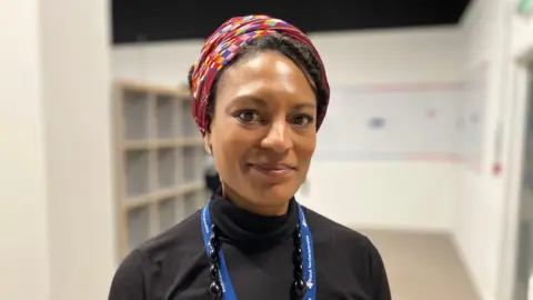 Sam Read/BBC A woman in a black roll neck jumper, a blue lanyard, and a multi-coloured hair cover smiles without teeth at the camera.