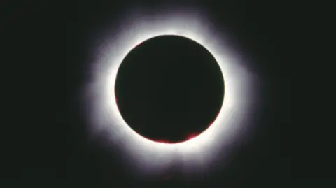 A total solar eclipse - a black circle with a glowing white outline, in a dark sky. 