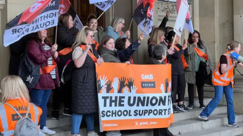 GMB protestors outside Birmingham City Council House. They are holding orange GMB-branded banners displaying slogans supporting  school support staff on strike.